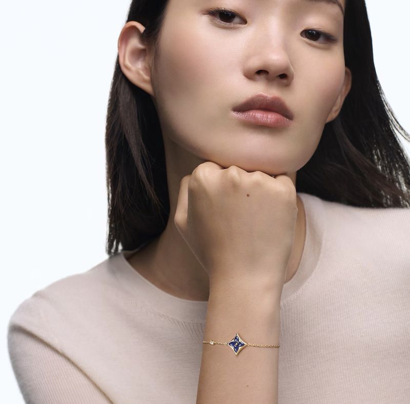 Close-up of Asian woman wearing a gold bracelet with a blue flower charm, set against a bright white background, jewellery advertisement.
