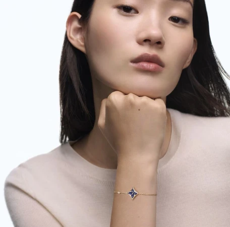 Close-up of Asian woman wearing a gold bracelet with a blue flower charm, set against a bright white background, jewellery advertisement.