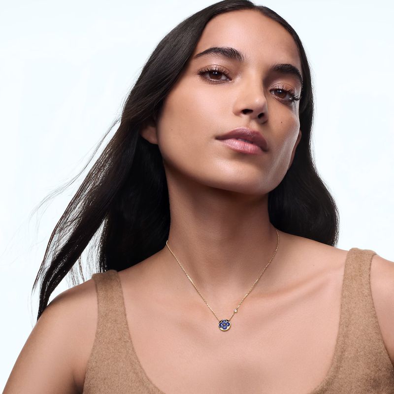 Close-up of an adult female model wearing a gold necklace with a round sapphire and diamond pendant in a studio with a soft light.