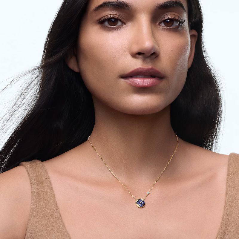 A woman with long dark hair wears a gold necklace with a blue and gold pendant against a bright background in a medium shot.