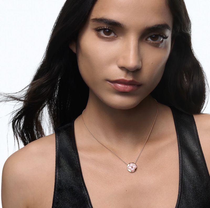 Close-up of a brunette woman wearing a flower pendant necklace and leather top against a white background in portrait orientation.