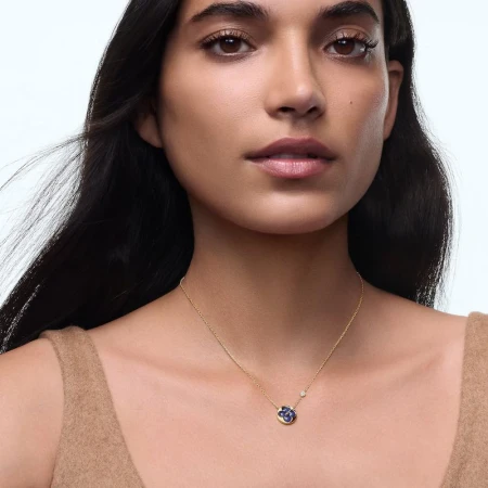 Close-up portrait of a woman wearing a gold necklace with a blue floral pendant against a clean white background.