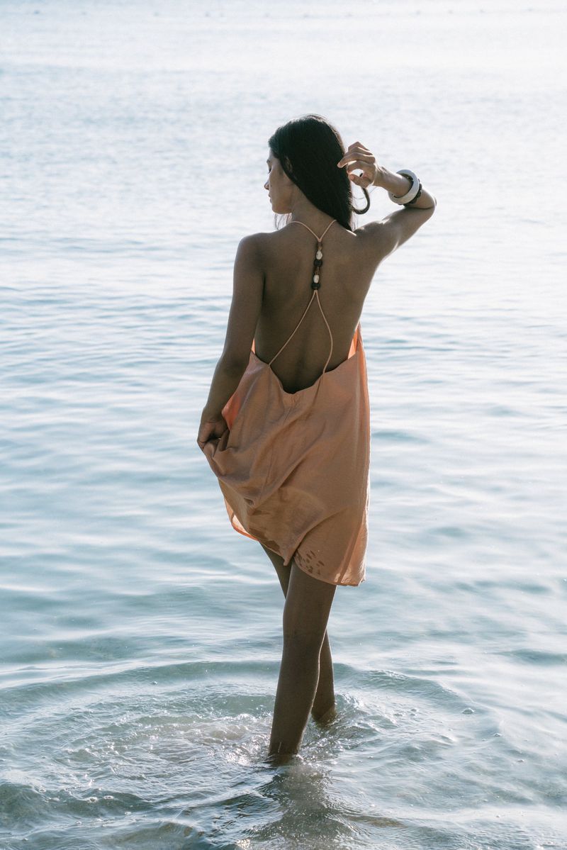 Woman in peach dress walking in shallow ocean water. Back view of adult female touching hair at the beach during the daytime.