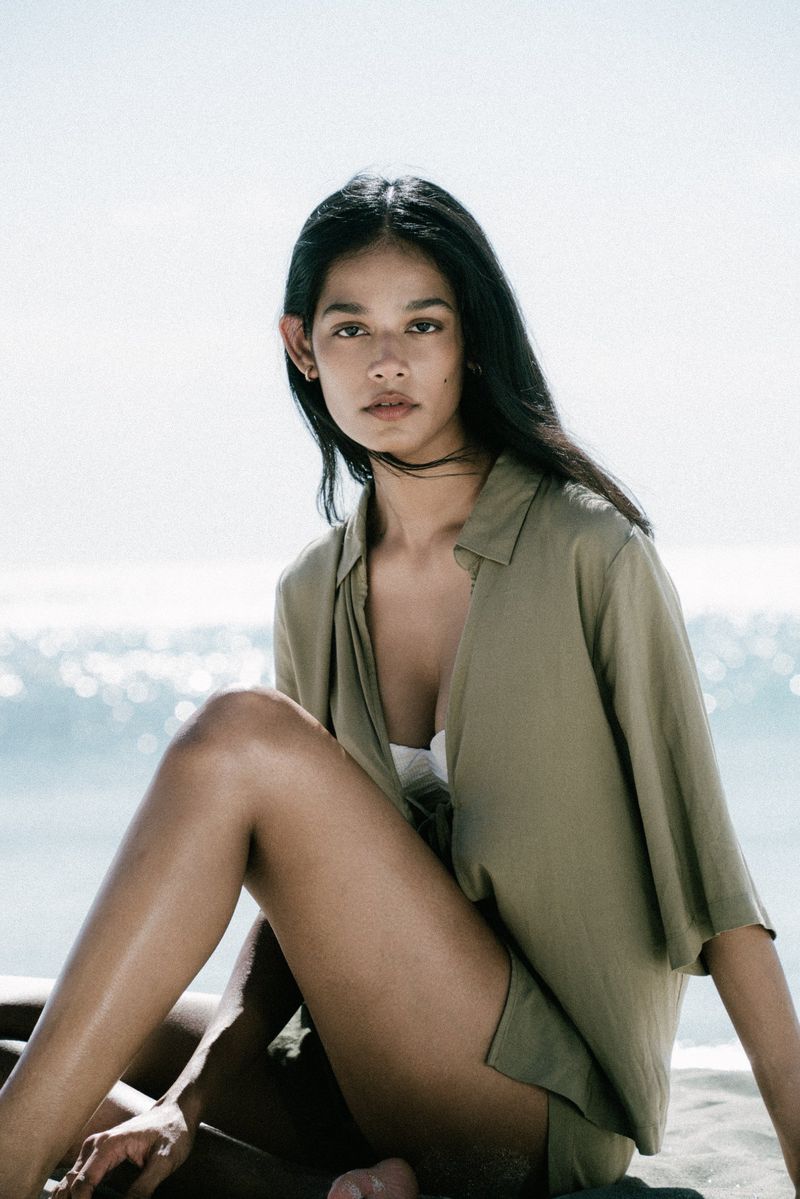 South Asian woman wearing an olive green button-down shirt sitting on a sandy beach by the ocean