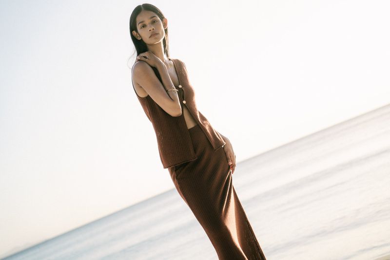 Brunette woman poses in a brown ribbed knit sleeveless top and skirt on a beach with light sky and water in the background.