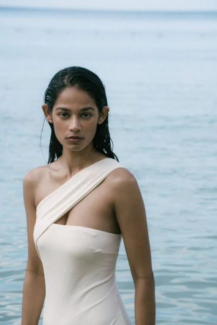 A woman in a swimsuit stands in shallow water; portrait shows sleek dark hair, cream-colored one-piece and a serene expression.