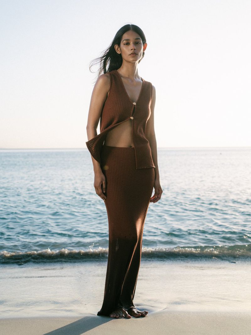 Woman in knit two-piece set poses barefoot on beach at water's edge in fashion shot with bright, diffused natural lighting. 