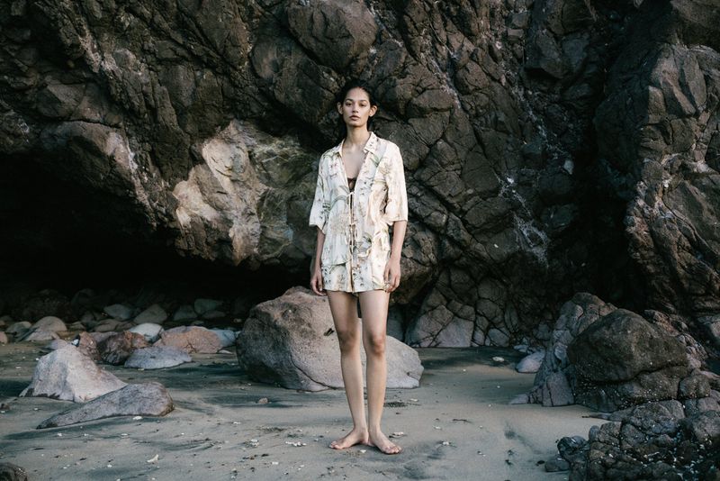 An adult female stands on a beach in a floral shirt and shorts, with large dark rocks in the background on an overcast day.