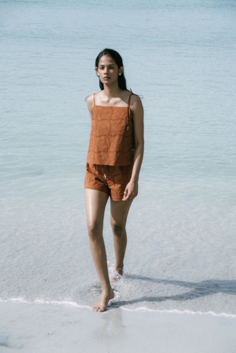 Young woman in a rust-colored linen top and shorts walking barefoot through shallow water at a tropical beach on a sunny day.