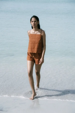 Young woman in a rust-colored linen top and shorts walking barefoot through shallow water at a tropical beach on a sunny day.