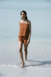 Model in rust-coloured two-piece set walking through shallow ocean water on a sandy beach