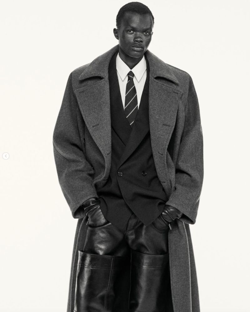 An African American male model poses in a grey coat, black leather pants, and gloves against a bright, neutral backdrop for a fashion shot.