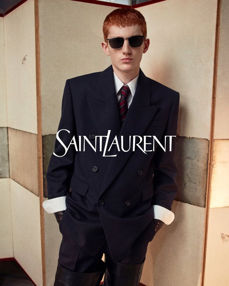 Male fashion model in oversized navy blazer and sunglasses posing in a studio with a neutral-colored panel backdrop; Saint Laurent logo.
