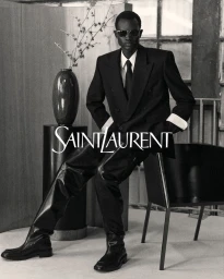 Black man wearing sunglasses and a black double-breasted suit seated in a studio setting for Saint Laurent.