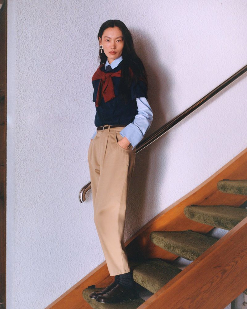 Stylish woman poses on stairs in a navy bow sweater, light blue shirt, tan pleated pants, black socks, and black shoes in an indoor setting.