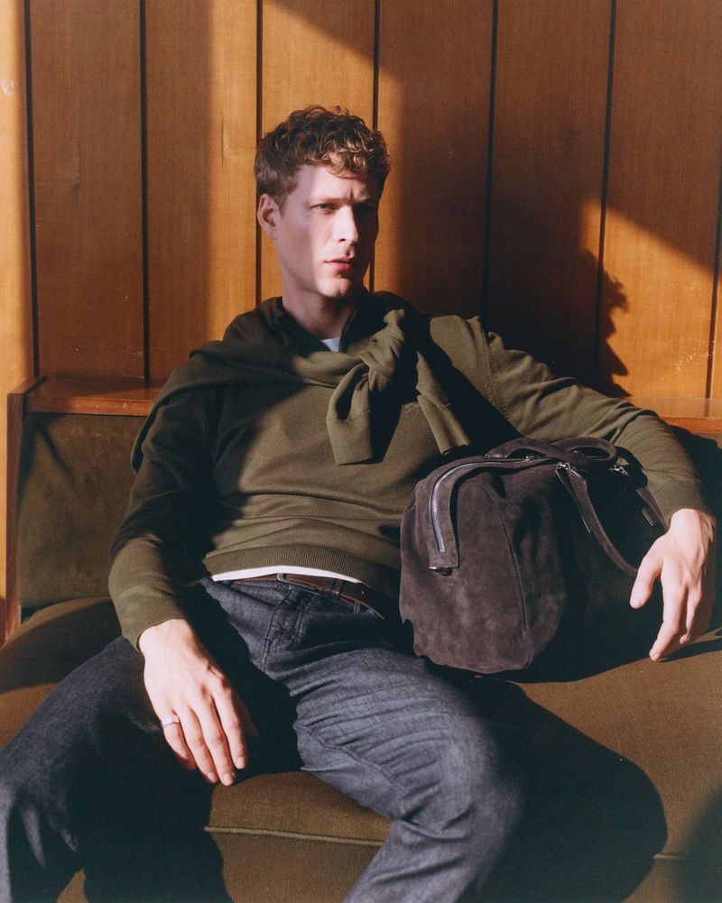 Adult male seated on olive-green sofa with wood paneling background, wearing casual clothing, and a dark duffel bag on his lap.