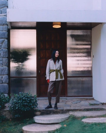 A woman stands outside a modern home, wearing a quilted jacket with a green belt, brown pants, and black shoes, posing on stone steps.