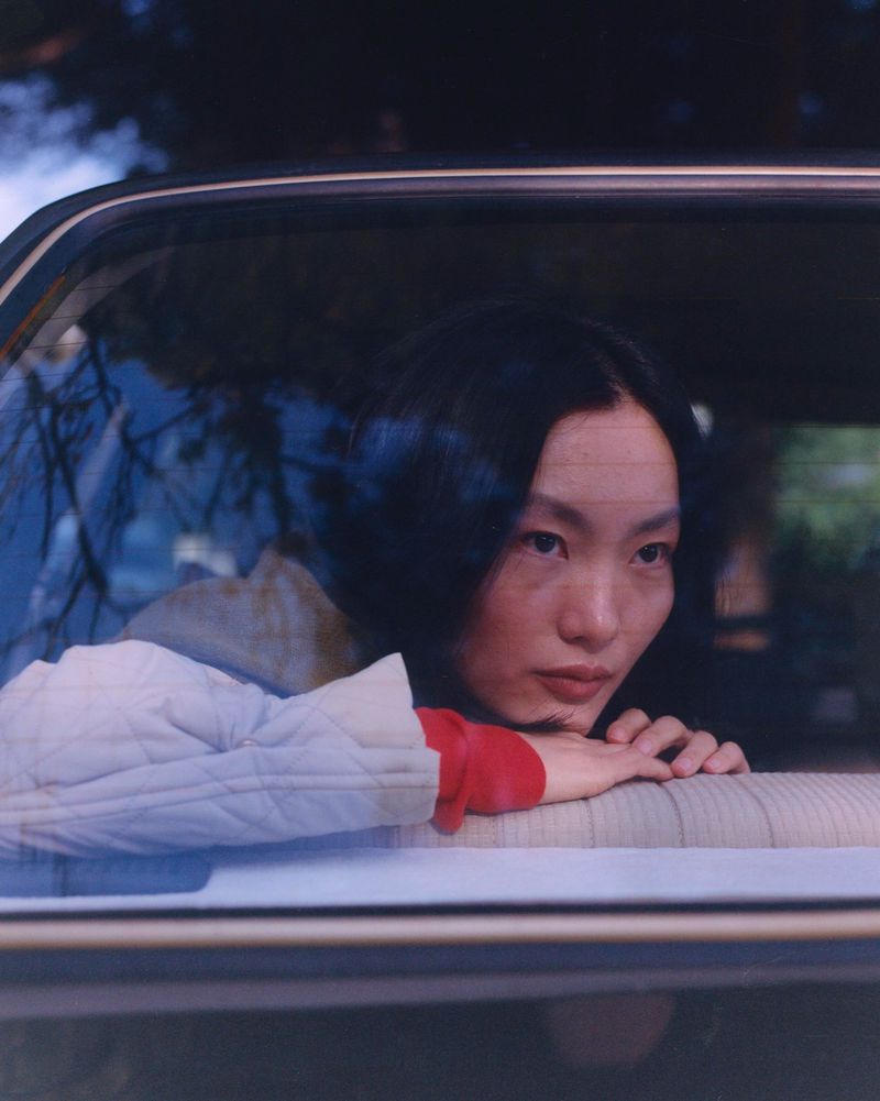 Portrait of a young Asian woman looking out of a car window, wearing a quilted jacket and red shirt, with a blurred background.