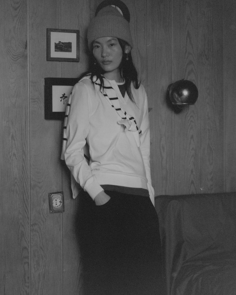 Monochrome medium shot of an Asian woman wearing a beanie, sweater, and dark pants, leaning against a wood-paneled wall indoors.