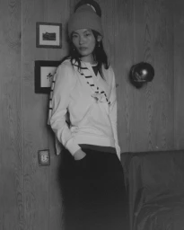 Model wearing a beanie and white sweater posing against a wood-paneled wall indoors