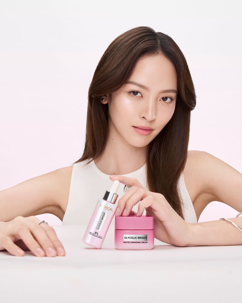 Cosmetic product promotion featuring a woman with long brown hair holding L'Oreal Glycolic-Bright serum and a jar of face cream.