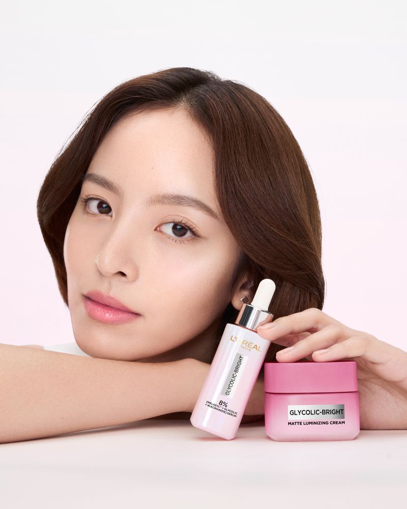 Asian woman poses with L'Oreal Glycolic-Bright skincare products: a serum bottle with dropper and a matte cream jar on a white surface.