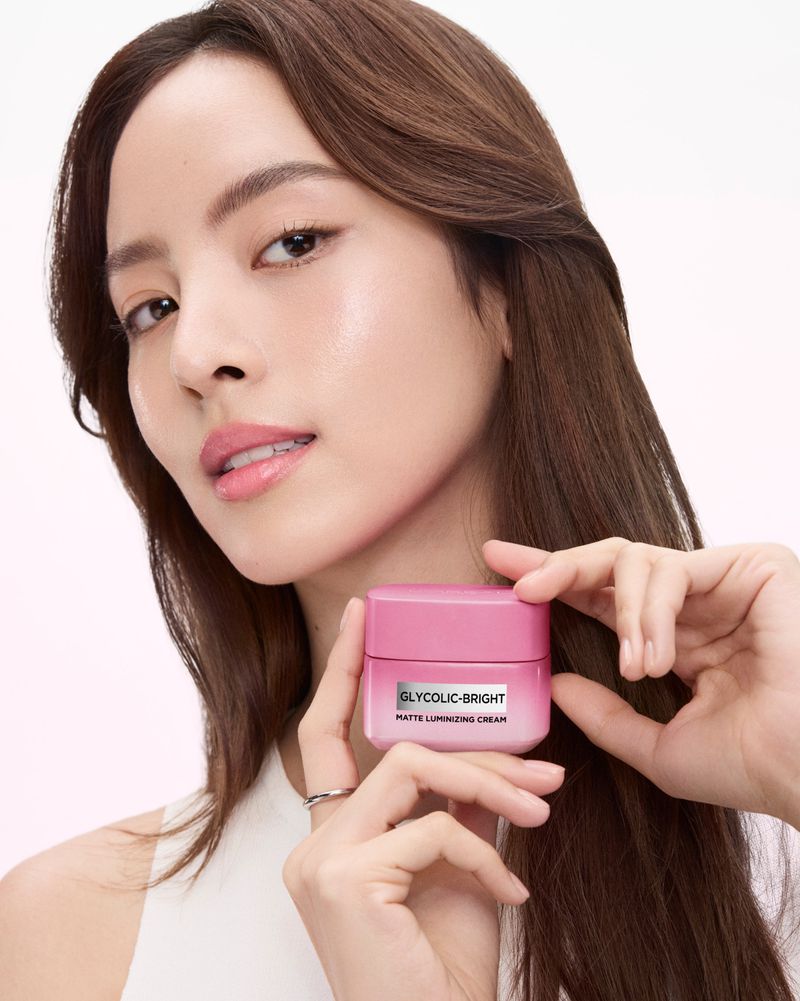 Asian adult female poses with a pink jar of Glycolic-Bright Matte Luminescing Cream, beauty shot for skincare, light background.