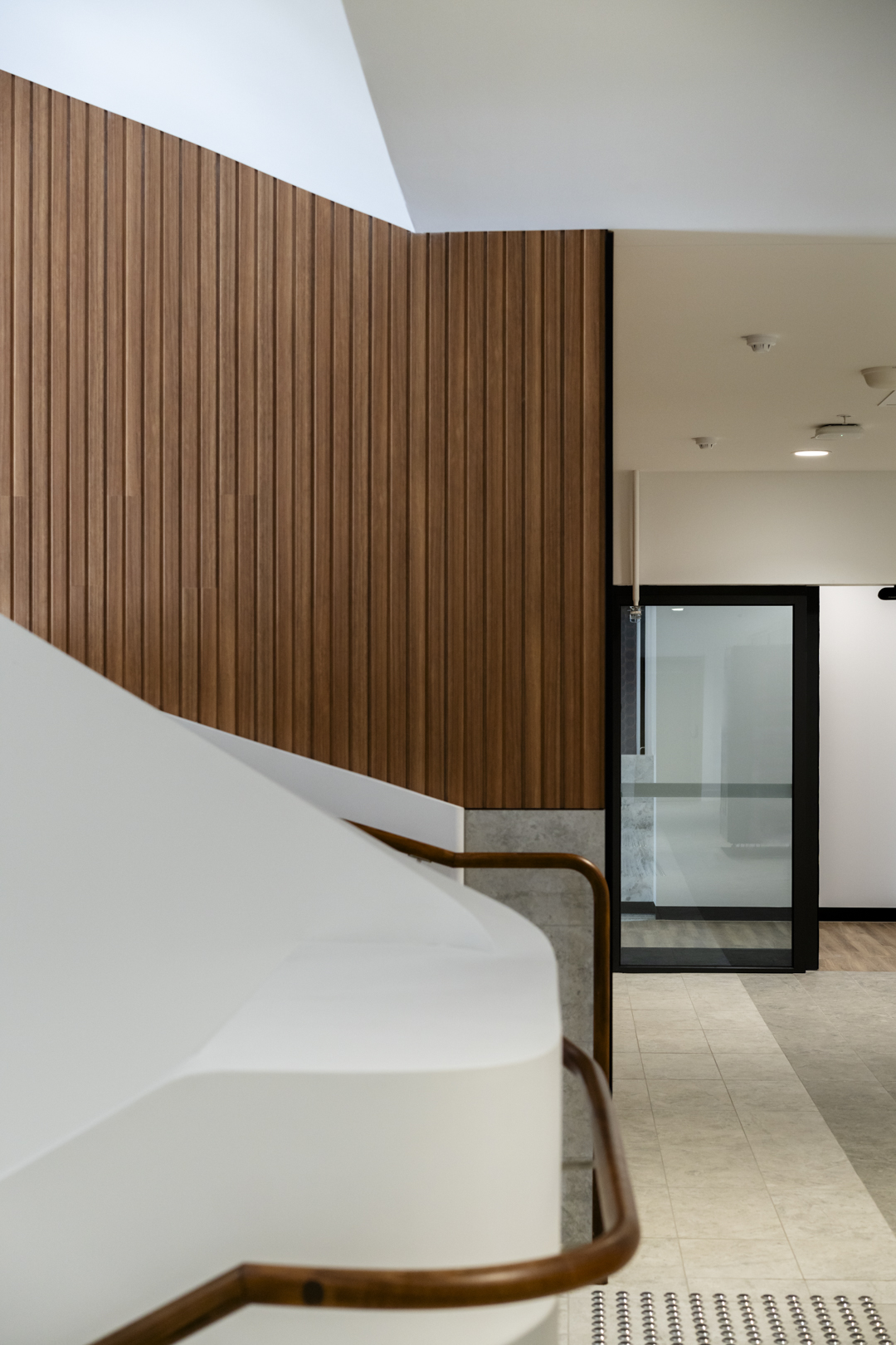 Indoor architectural shot featuring a modern white staircase with brown railing, set against a wooden panel wall and opening to a doorway.
