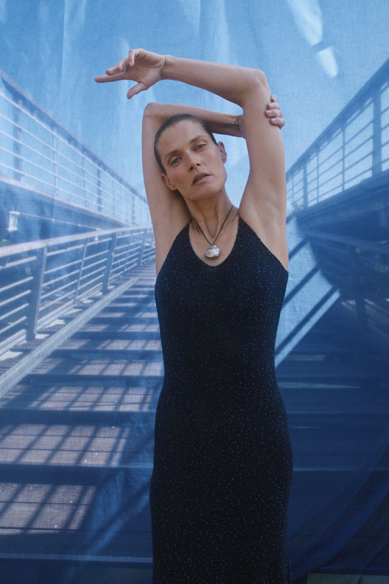 Elegant woman in a black dress poses against a blue backdrop with a bridge graphic, arms raised, showcasing fashion and style.