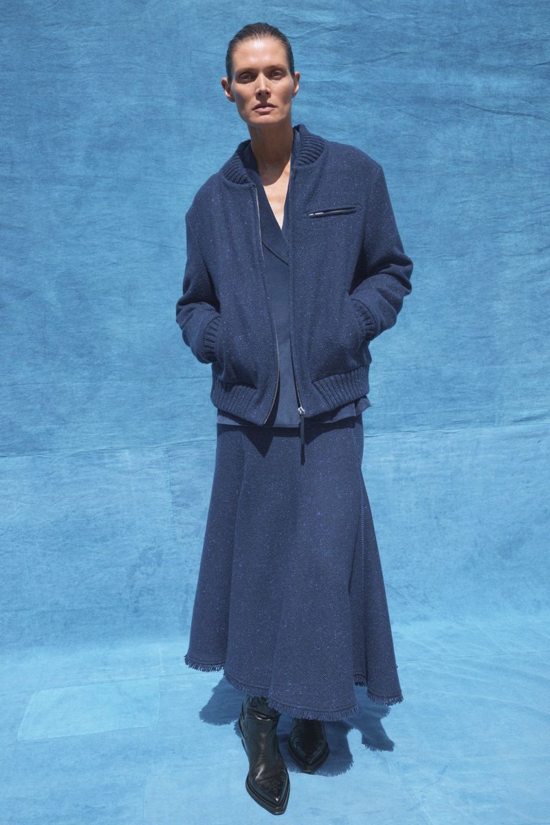 A slender adult woman poses in blue knitwear against a blue fabric background, wearing a cardigan jacket, skirt, and shiny black heeled boots.