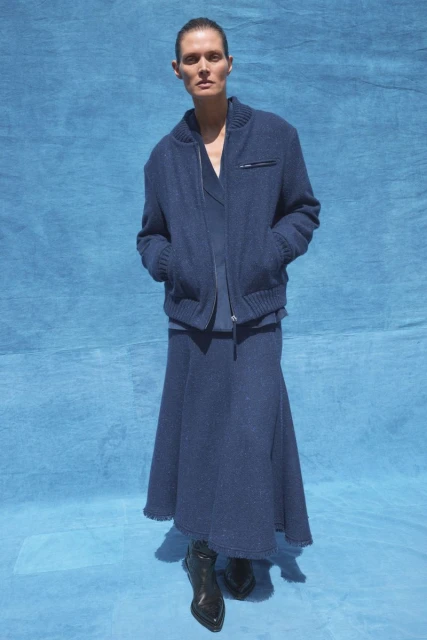 A slender adult woman poses in blue knitwear against a blue fabric background, wearing a cardigan jacket, skirt, and shiny black heeled boots.