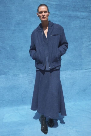 A slender adult woman poses in blue knitwear against a blue fabric background, wearing a cardigan jacket, skirt, and shiny black heeled boots.