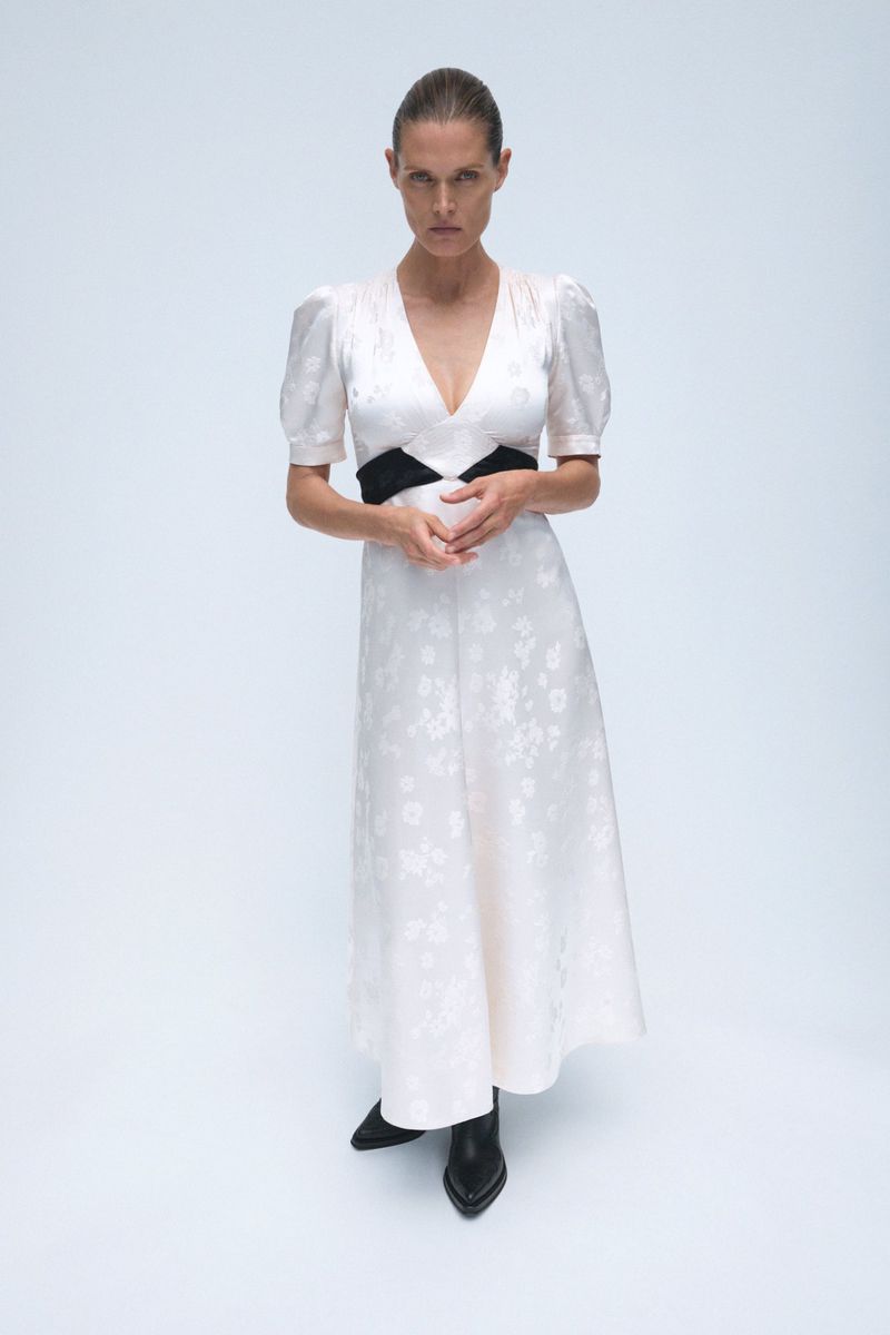 Full-length studio shot of a slim adult woman in a light floral dress with a black waistband and black boots, standing against a white backdrop.
