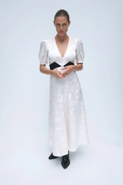Full-length studio shot of a slim adult woman in a light floral dress with a black waistband and black boots, standing against a white backdrop.