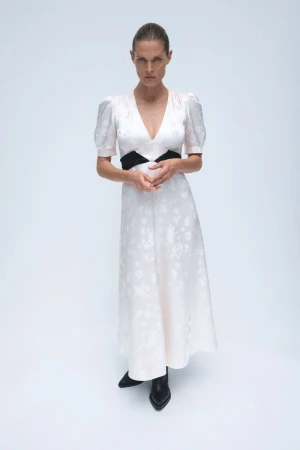 Full-length studio shot of a slim adult woman in a light floral dress with a black waistband and black boots, standing against a white backdrop.