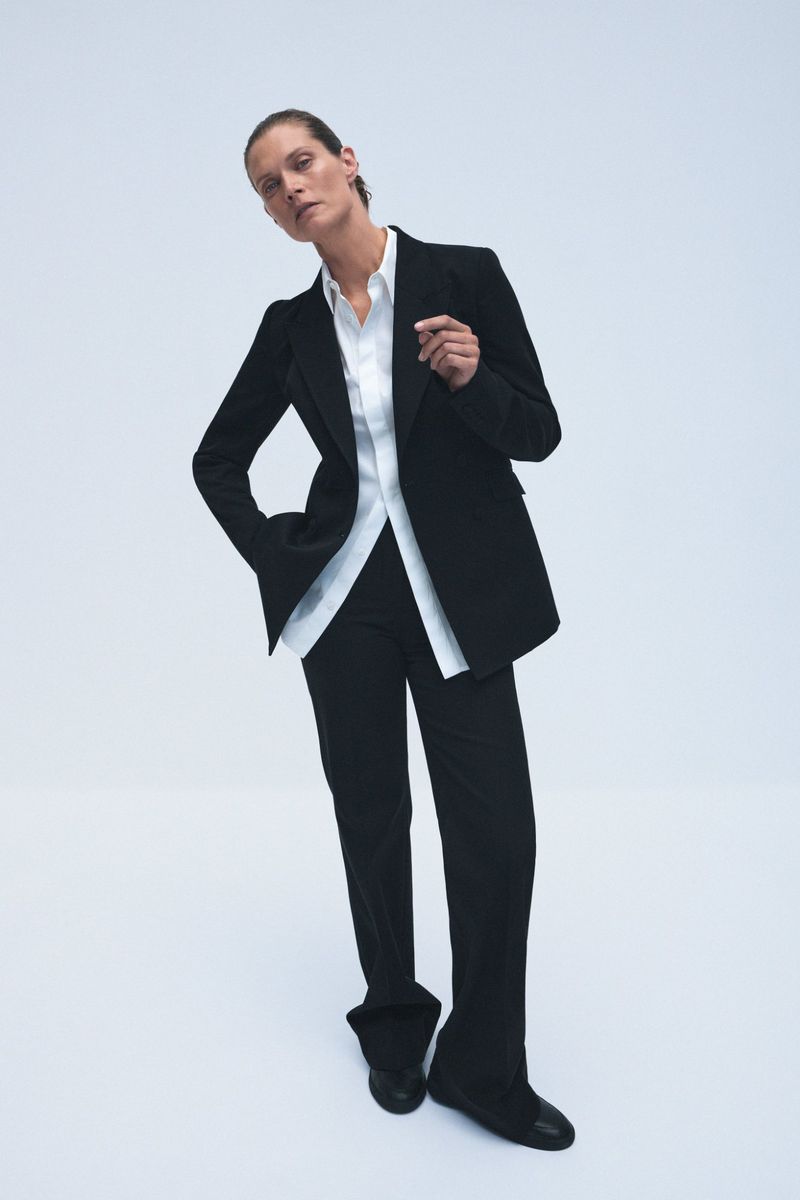 Elegant adult female posing in a black suit and white button-down shirt against a bright white backdrop in a fashion setting.