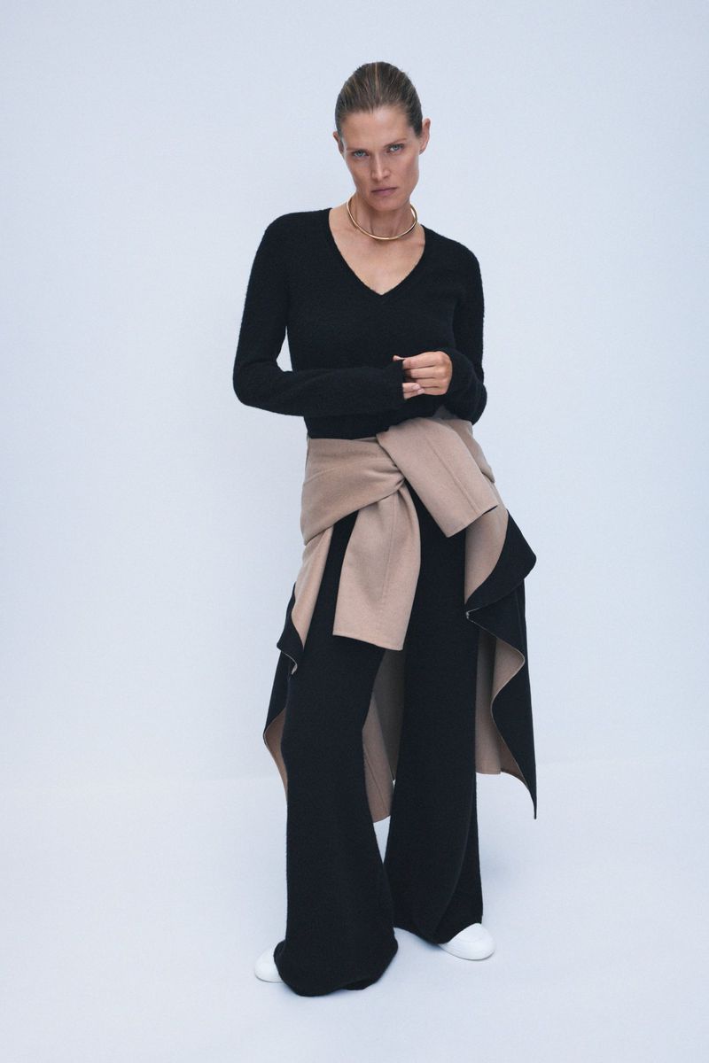 Fashionable adult woman in a v-neck sweater and wide-leg pants strikes a confident pose in a studio with soft, neutral lighting.