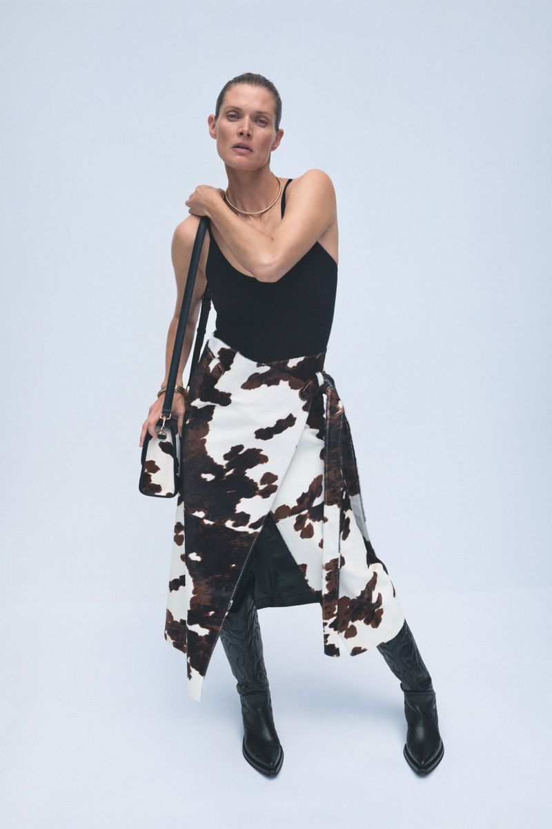 A woman poses in a black tank top, cow-print wrap skirt, and black heeled boots, holding a matching bag, against a white backdrop.