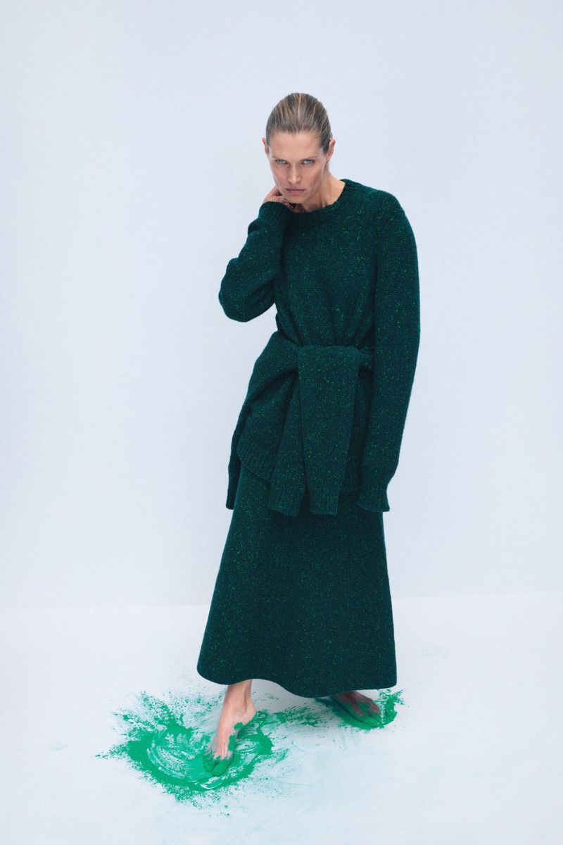 Elegant woman in a long-sleeved green sweater dress poses in a studio, standing barefoot on a splash of green powder paint.