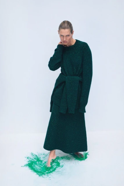 Elegant woman in a long-sleeved green sweater dress poses in a studio, standing barefoot on a splash of green powder paint.