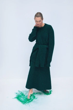 Elegant woman in a long-sleeved green sweater dress poses in a studio, standing barefoot on a splash of green powder paint.