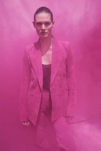 Woman poses in pink pantsuit against smoky pink background in medium shot. Fashion editorial, studio shot, and monochromatic palette.