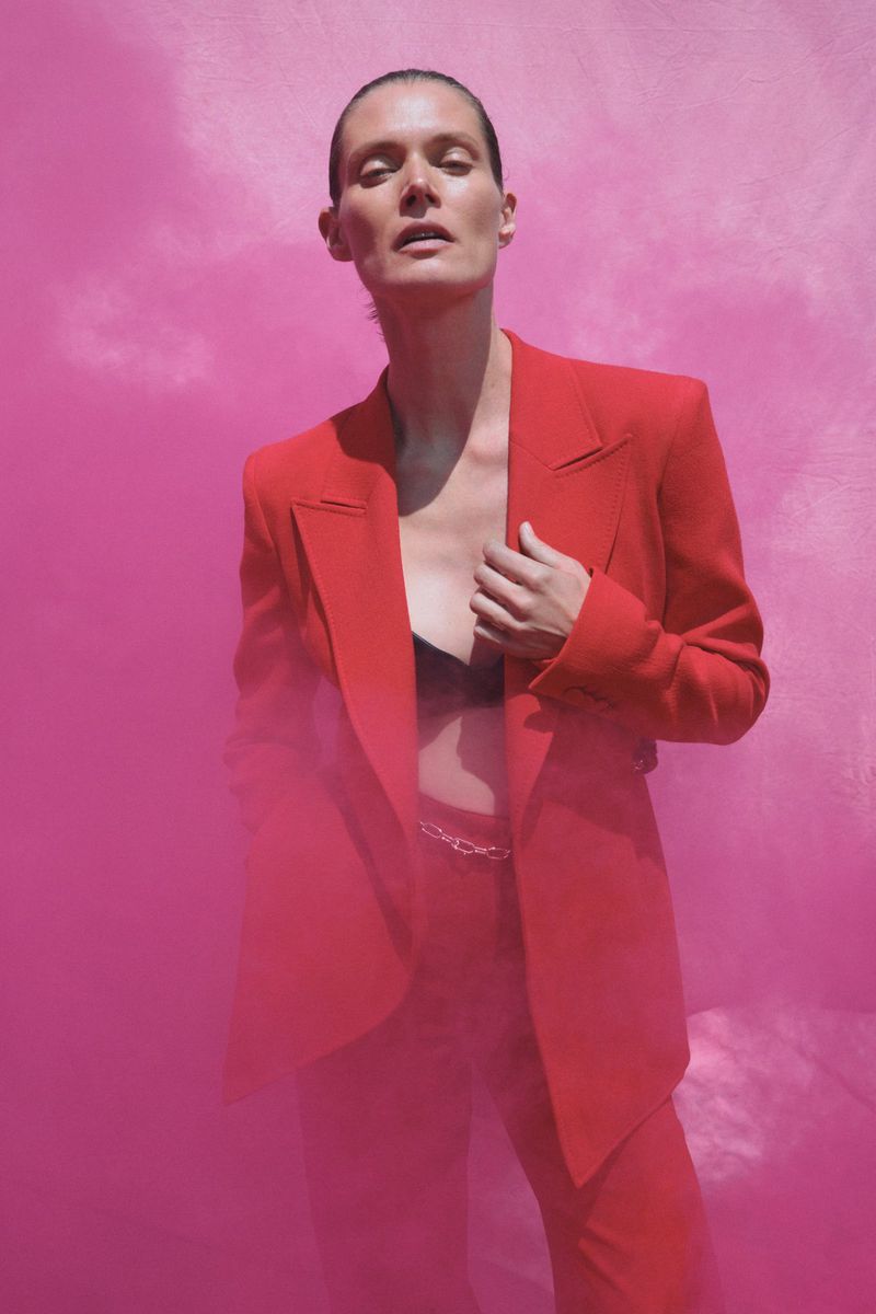 Adult female fashion model poses in a red suit against a pink background, smoke rising in front of her.