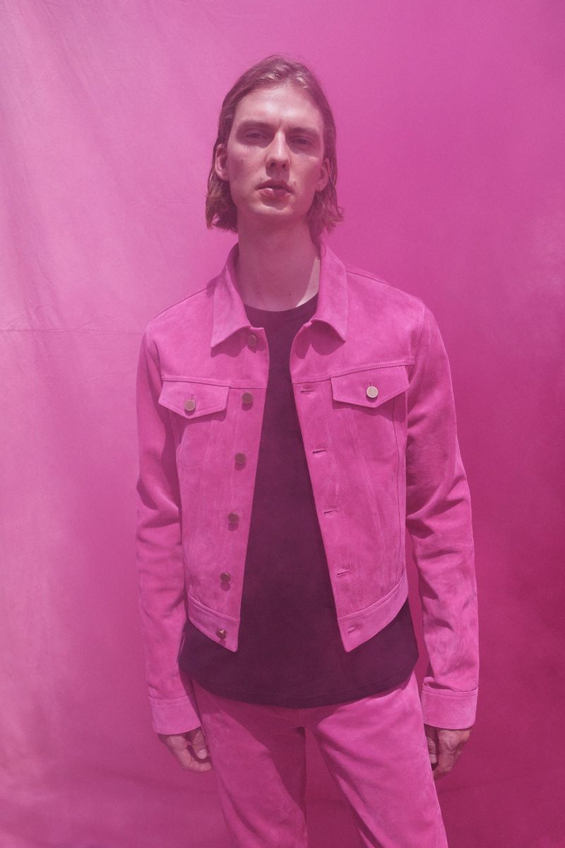 Man with long hair wearing a pink suede jacket and pants poses in front of a pink background in a fashion or editorial setting.