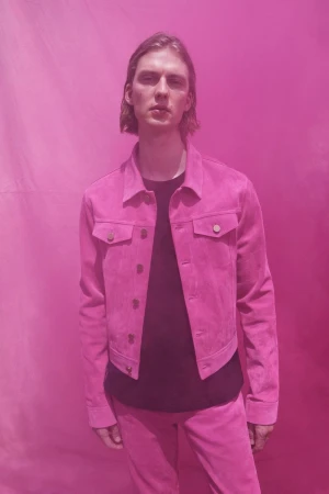 Man with long hair wearing a pink suede jacket and pants poses in front of a pink background in a fashion or editorial setting.