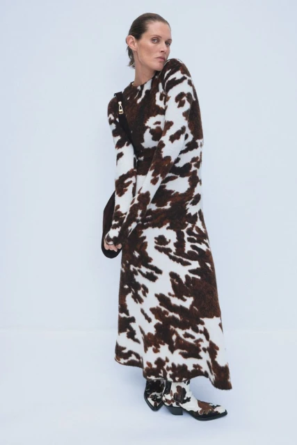 Adult female poses in a brown and white cow-print dress and matching boots against a clean white backdrop in a studio fashion shot.