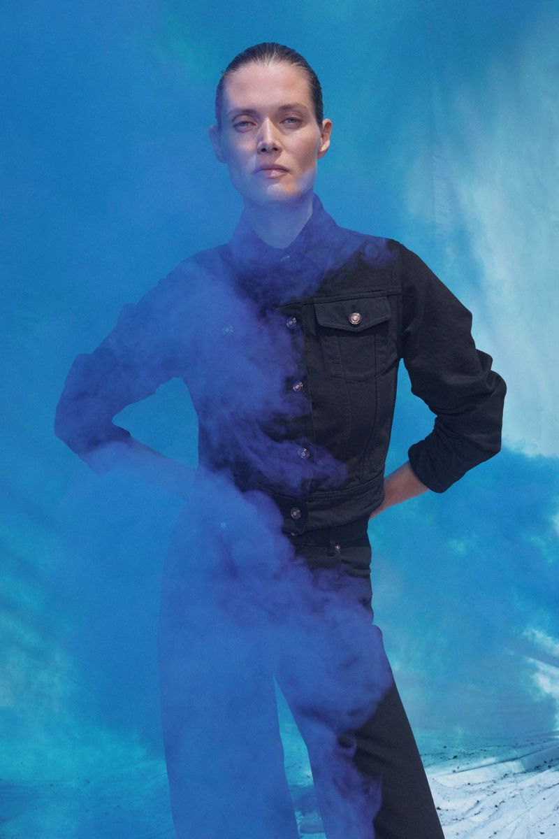 An adult female stands against a bright blue background with hands on her hips, partially obscured by a cloud of deep blue smoke.