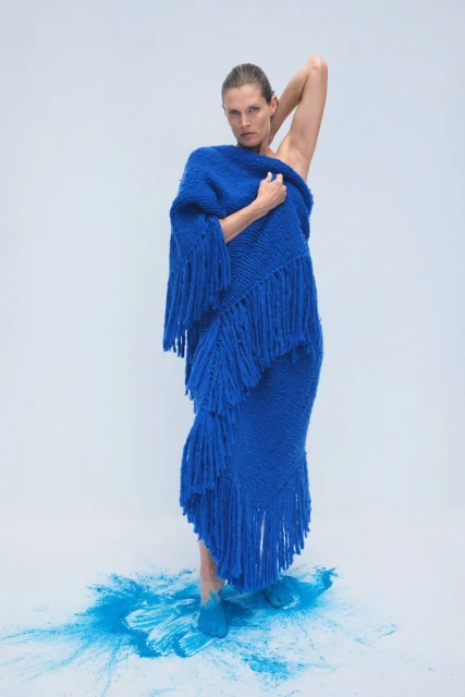 A model poses in a photo studio wearing a blue knit poncho dress with a fringe, creating a fashion-forward and visually striking image.
