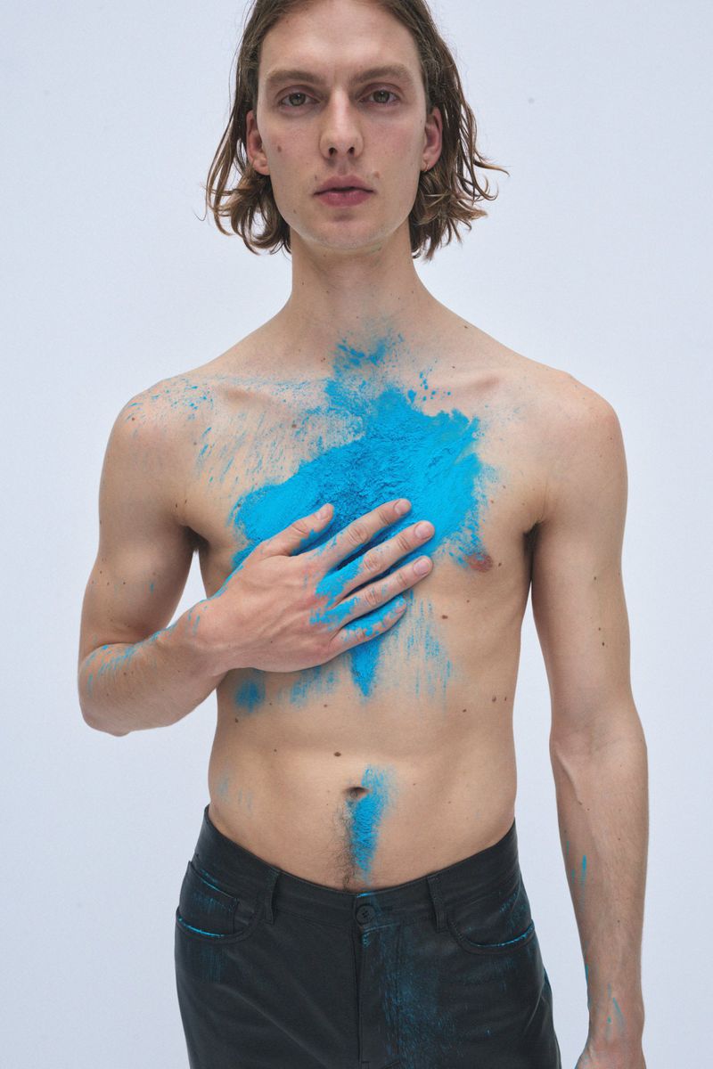 Nude male model in dark pants posing with blue powder pigment on chest, holding his hand to his chest, against a plain white backdrop.