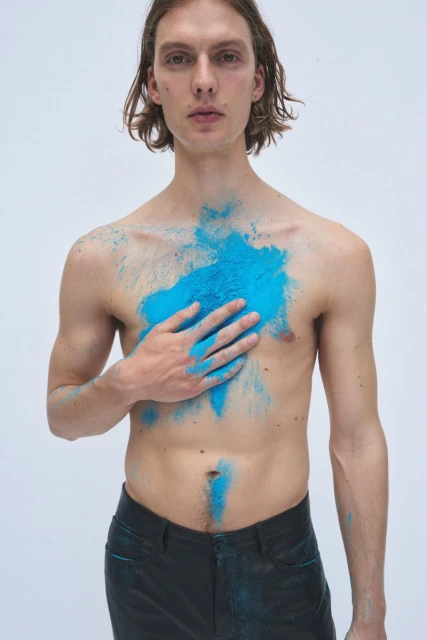 Nude male model in dark pants posing with blue powder pigment on chest, holding his hand to his chest, against a plain white backdrop.